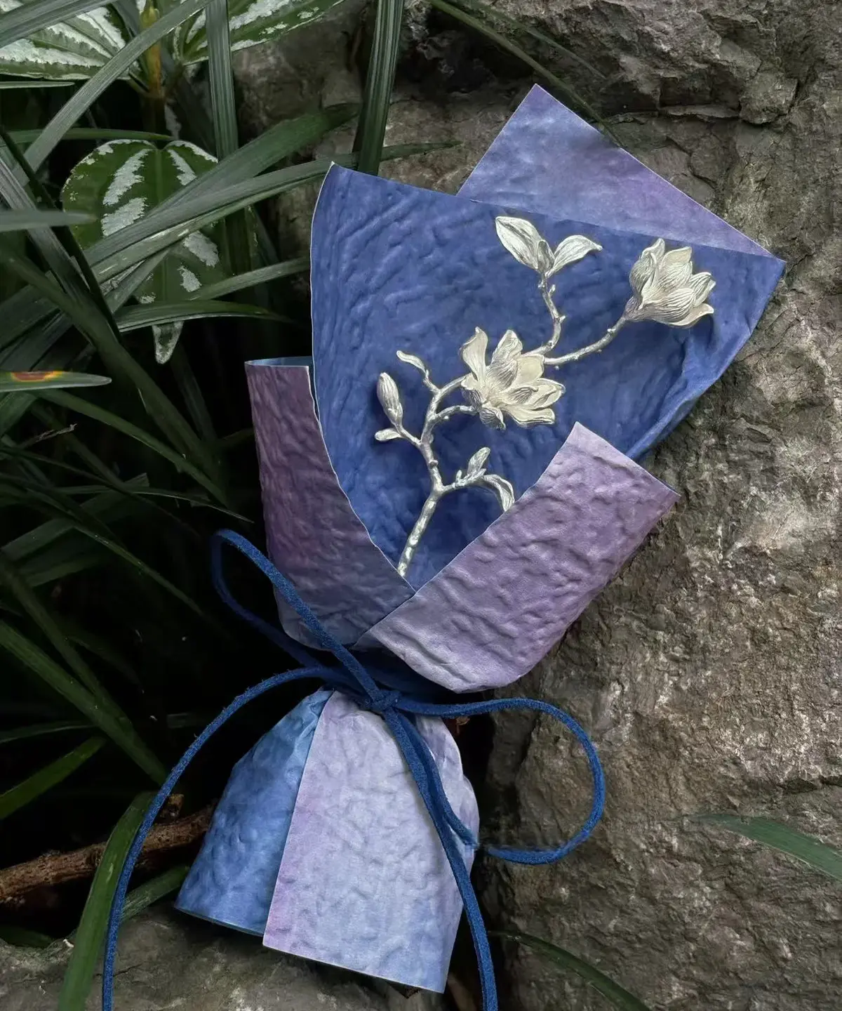 925 Sterling Silver Magnolia Bouquet wrapped in gradient blue and purple paper with silver floral branch, tied with blue ribbon; a nature-inspired Handmade Art Gift styled against rocks and green foliage.