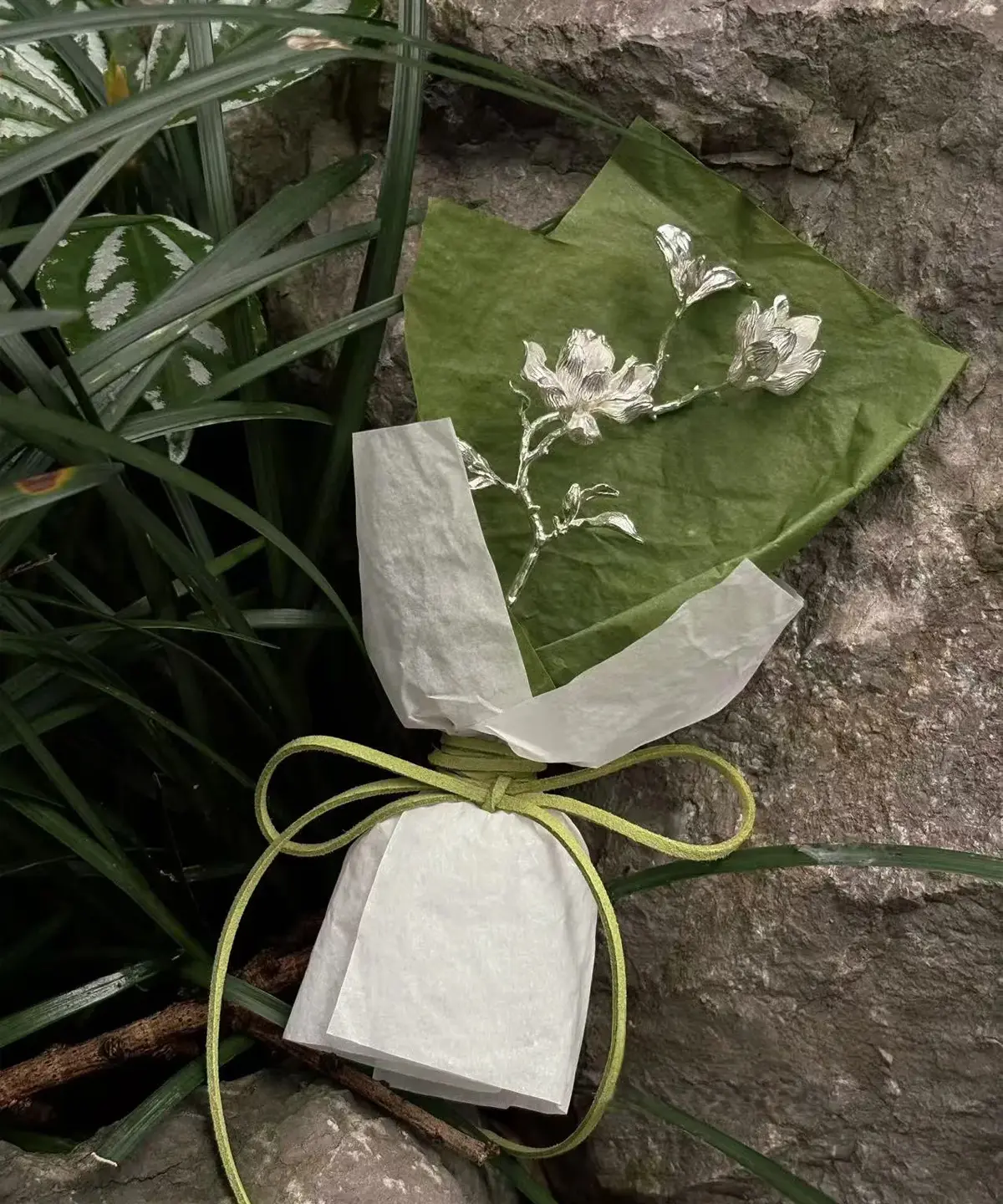 925 Sterling Silver Magnolia Bouquet wrapped in green and white paper with silver floral branch, tied with green string; a nature-inspired Handmade Art Gift styled against rocks and foliage.