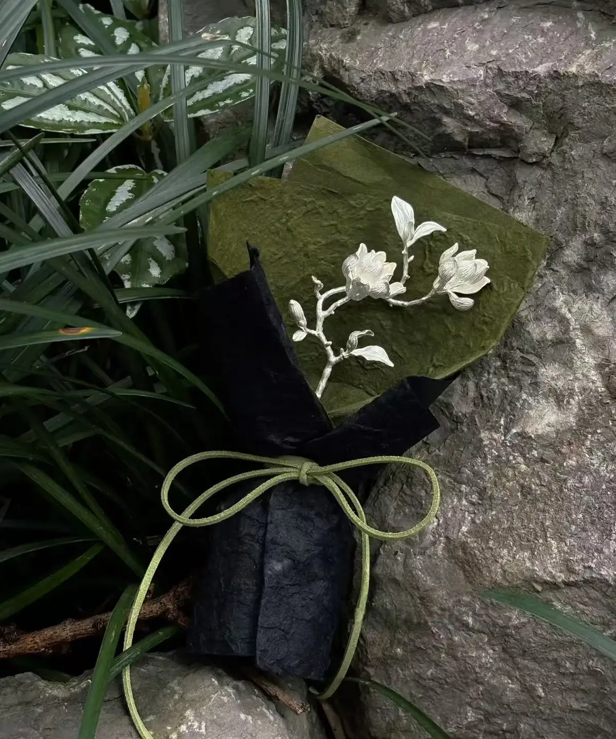 925 Sterling Silver Magnolia Bouquet wrapped in black and green paper with green string, featuring sculptural silver magnolia flower and leaves; a nature-inspired Handmade Art Gift styled among rocks and foliage.