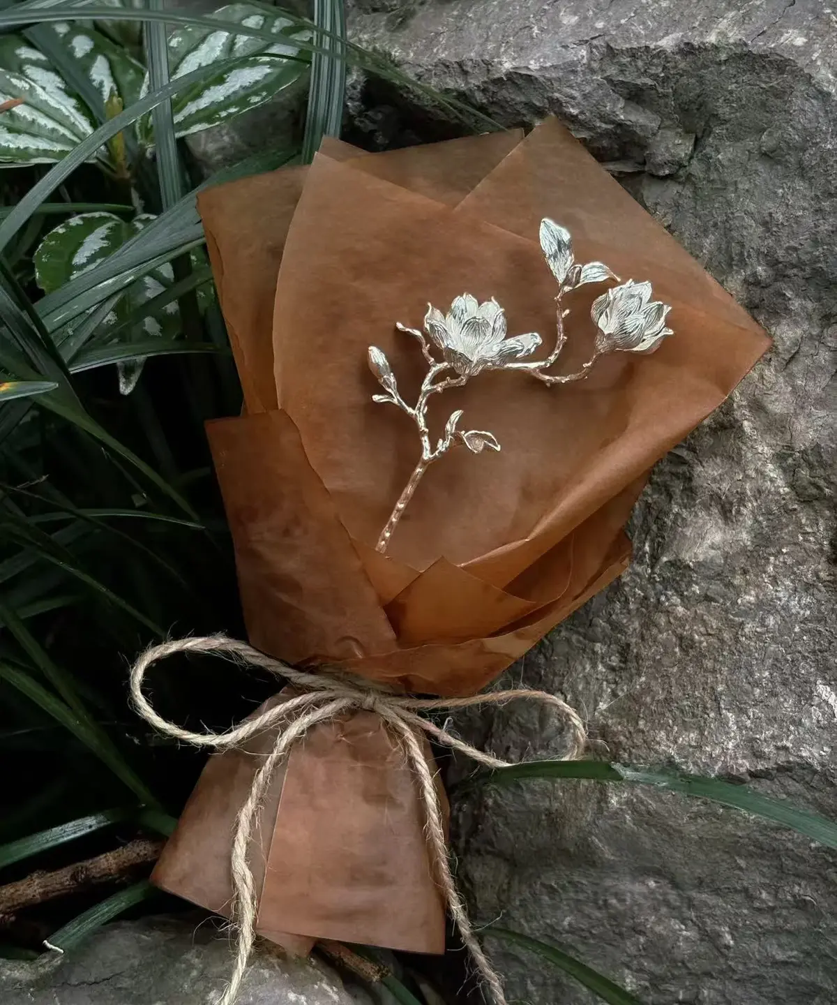 925 Sterling Silver Magnolia Bouquet wrapped in brown parchment paper and tied with rustic twine, featuring sculptural silver magnolia blossoms; a nature-inspired Handmade Art Gift styled against stone and green foliage.