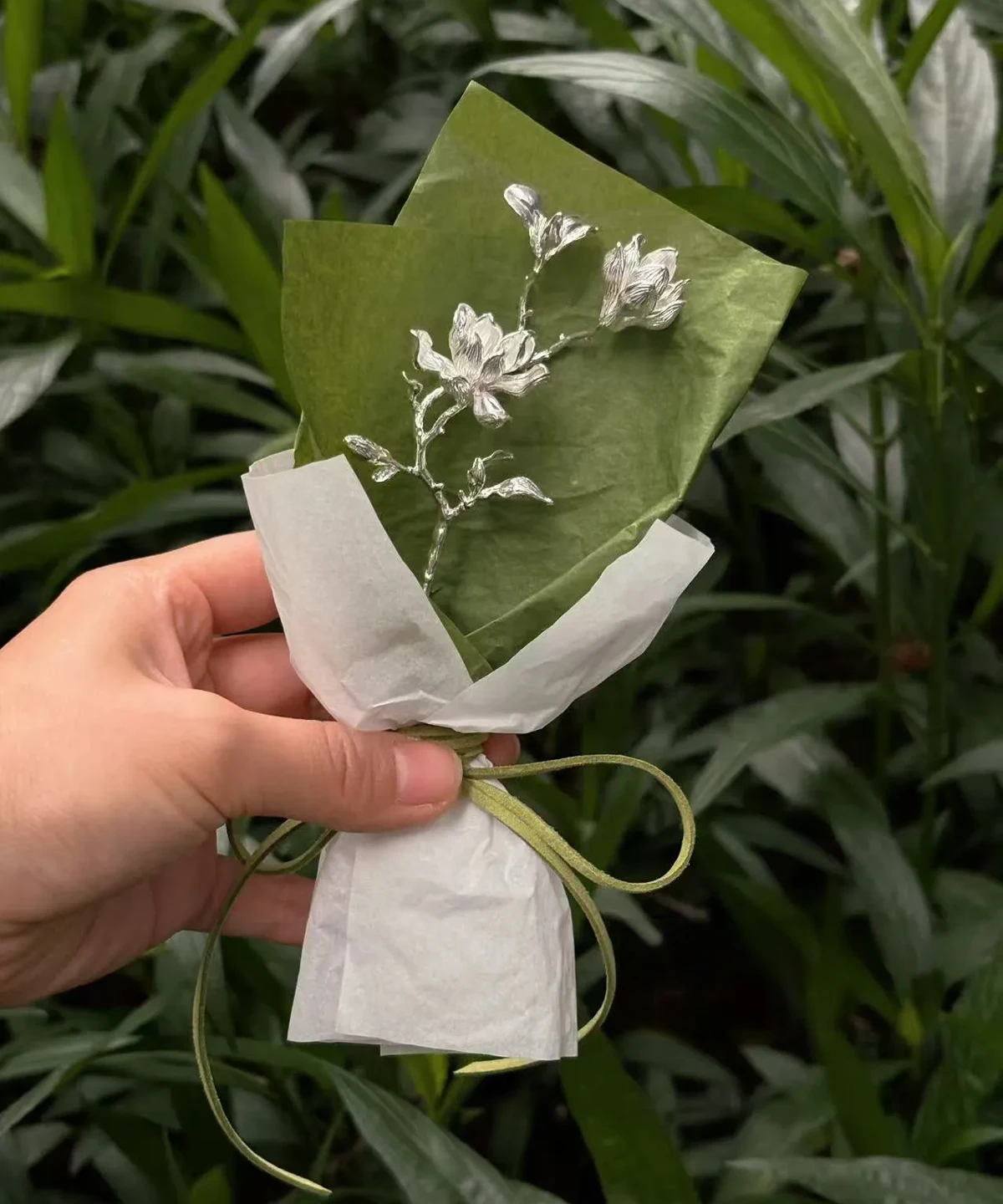 Hand holding a 925 Sterling Silver Magnolia Bouquet wrapped in green and white paper with green ribbon; a botanical Handmade Art Gift featuring sculptural silver magnolia flowers, styled against lush garden foliage.