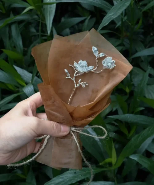 Hand holding a 925 Sterling Silver Magnolia Bouquet wrapped in brown paper and tied with rustic string; a botanical Handmade Art Gift featuring sculptural silver magnolia blossoms, styled against lush green foliage and stone.