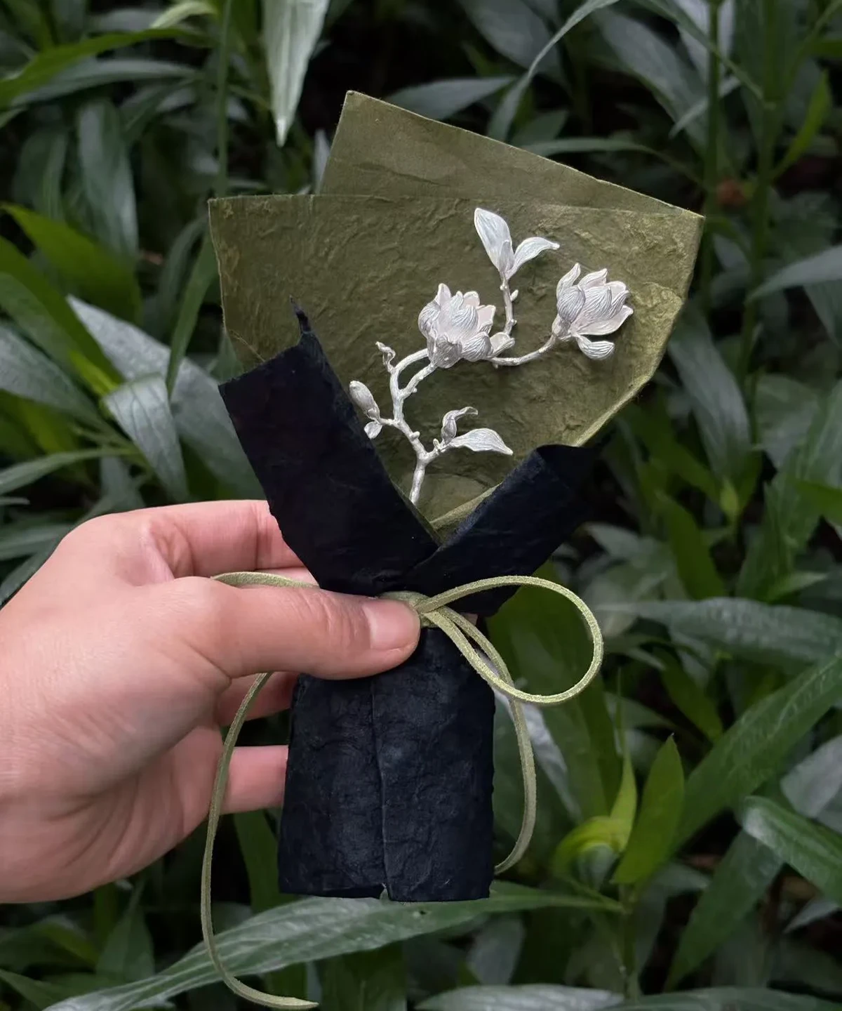 Hand holding a 925 Sterling Silver Magnolia Bouquet wrapped in green and black paper with green string; a botanical Handmade Art Gift featuring sculptural silver magnolia flowers and leaves, styled against lush garden foliage.