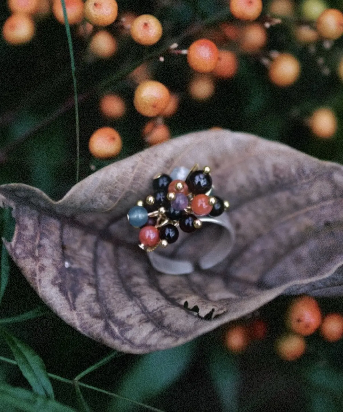 925 Sterling Silver Ring with multicolored Natural Stone beads in black, red, blue, and purple, accented with golden spheres and leaf charm; a handcrafted Silver Ring displayed on dried leaf with berry plant background.