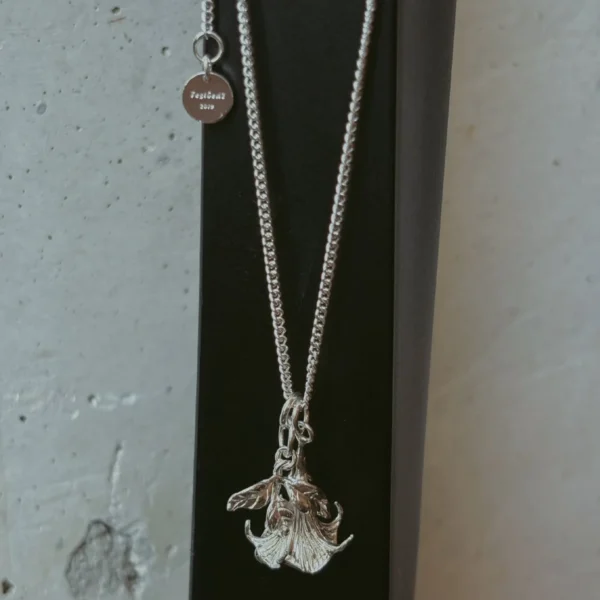 Handmade S925 Silver Necklace featuring a detailed datura flower pendant and engraved custom disc tag “Festbois 2019”; a botanical-inspired Flower Necklace displayed on black stand with textured concrete background.