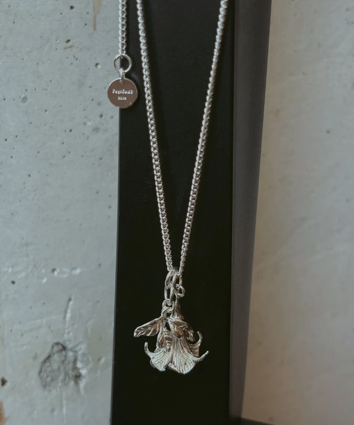 Datura Flower Necklace-Handmade S925 Silver With Custom Disc 02 Handmade S925 Silver Necklace featuring a detailed datura flower pendant and engraved custom disc tag “Festbois 2019”; a botanical-inspired Flower Necklace displayed on black stand with textured concrete background.
