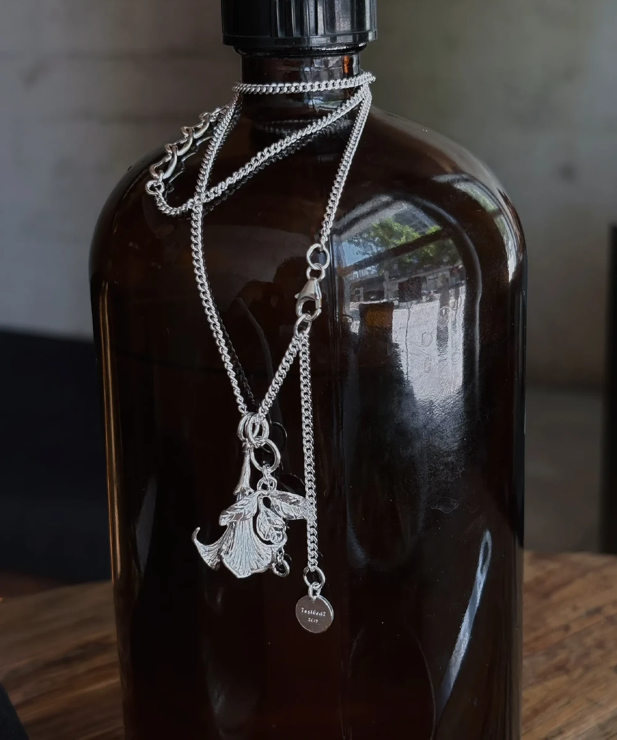 Datura Flower Necklace-Handmade S925 Silver With Custom Disc 03 Handmade S925 Silver Necklace with detailed datura flower pendant and custom disc tag engraved “Tsubasa 925”; a botanical-inspired Flower Necklace draped around a dark glass bottle, highlighting silver craftsmanship and contrast.