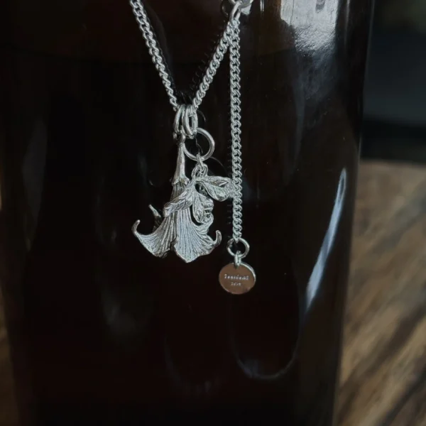 Handmade S925 Silver Necklace with detailed datura flower pendant and custom disc tag, featuring petal and leaf engravings; a unique Flower Necklace displayed against a dark glossy surface for contrast and elegance.