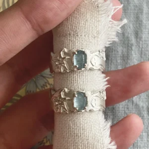Two Handmade 999 Silver Rings with rectangular Aquamarine gemstones, held between fingers on frayed white fabric; intricate ocean-inspired designs with starfish and wave motifs highlight the elegance of this March birthstone Aquamarine Ring.