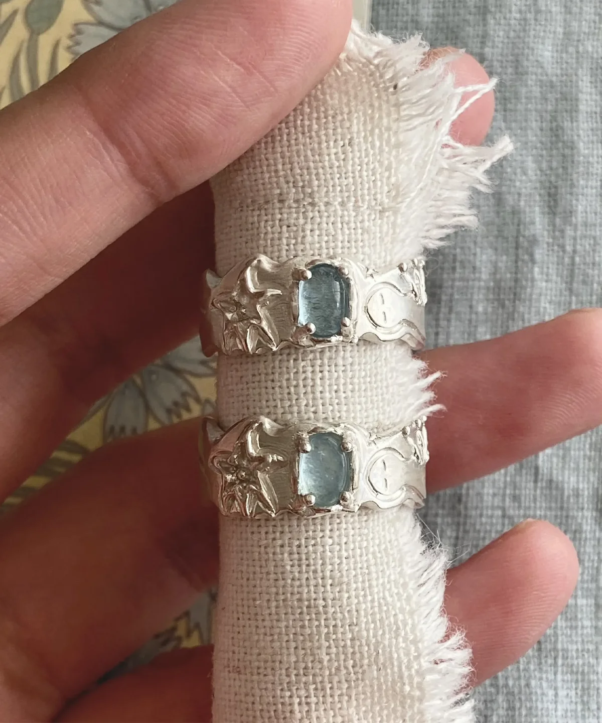 Two Handmade 999 Silver Rings with rectangular Aquamarine gemstones, held between fingers on frayed white fabric; intricate ocean-inspired designs with starfish and wave motifs highlight the elegance of this March birthstone Aquamarine Ring.