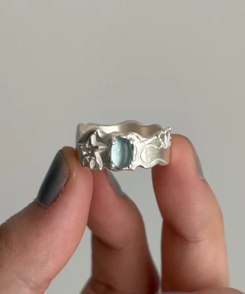 Hand holding a Handmade 999 Silver Ring with oval Aquamarine gemstone and marine-inspired motifs including starfish and waves; a poetic Aquamarine Ring celebrating March birthstone craftsmanship in sculptural 999 Silver Ring detail.