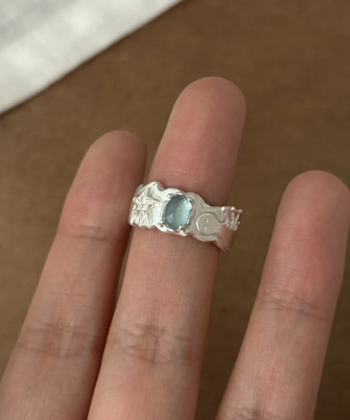 Close-up of a hand wearing a Handmade 999 Silver Ring with oval Aquamarine gemstone, featuring starfish, crescent moon, and wave motifs; a poetic Aquamarine Ring crafted in fine silver, celebrating March birthstone elegance and ocean-inspired artistry.