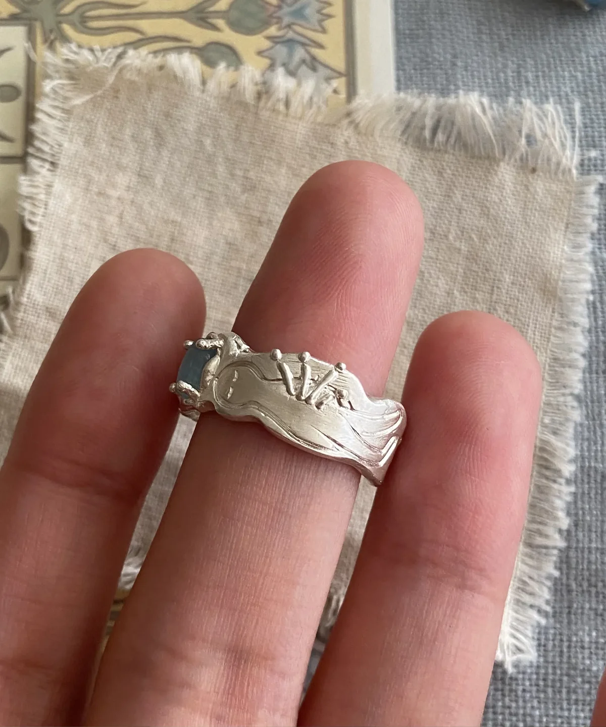 Hand wearing a Handmade 999 Silver Ring with sculptural reclining figure and a small Aquamarine gemstone; artistic design evokes mythic elegance and celebrates the March birthstone in finely crafted 999 Silver Ring detail.