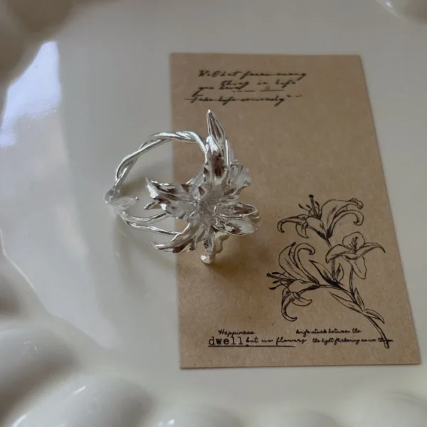 Handmade S925 Sterling Silver Lily Ring 06 Handmade S925 Sterling Silver Lily Ring with sculpted petals and vine-textured band, placed on a floral card in a ceramic dish; a botanical Silver Ring inspired by the elegance of the lily flower, blending nature and artistry.