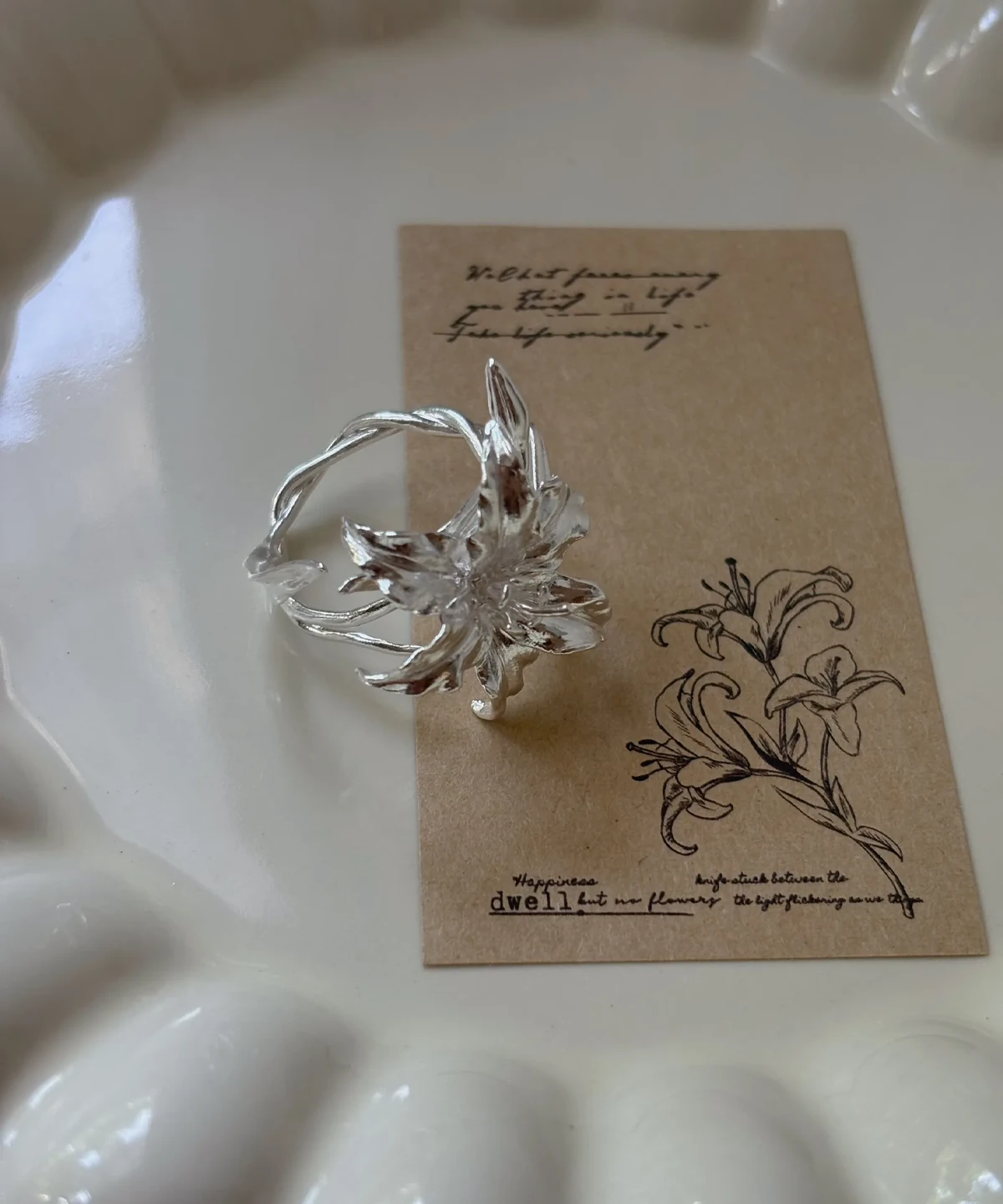 Handmade S925 Sterling Silver Lily Ring 06 Handmade S925 Sterling Silver Lily Ring with sculpted petals and vine-textured band, placed on a floral card in a ceramic dish; a botanical Silver Ring inspired by the elegance of the lily flower, blending nature and artistry.
