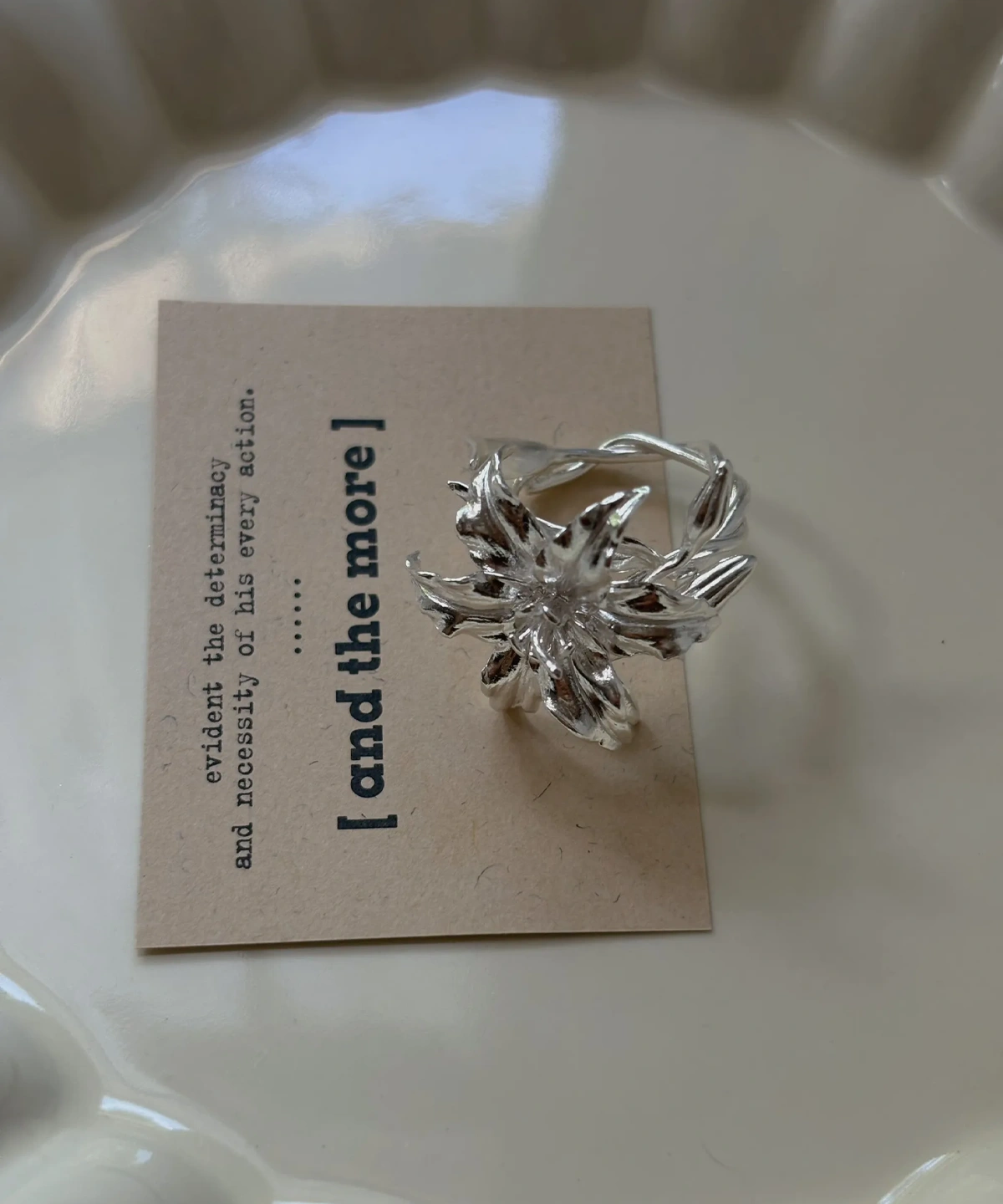 Handmade S925 Sterling Silver Lily Ring 07 Handmade S925 Sterling Silver Lily Ring with sculpted petals and vine-textured band, placed on a beige card in a ceramic dish; a botanical Silver Ring inspired by the lily flower’s elegance and crafted with artisanal detail.