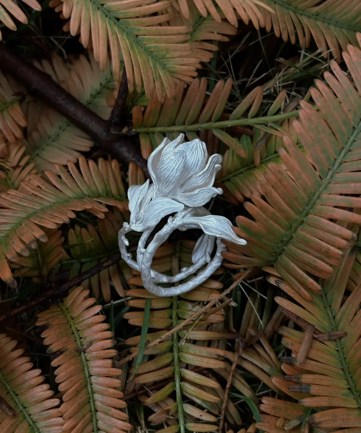 Handmade S925 Sterling Silver Magnolia Ring with sculpted petals and stem details, placed on dried fern leaves; a nature-inspired Silver Ring celebrating Magnolia Jewelry craftsmanship and botanical elegance.
