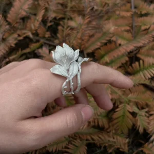 Handmade S925 Sterling Silver Magnolia Ring worn across two fingers, with sculpted petals and stems; a nature-inspired Silver Ring showcasing Magnolia Jewelry craftsmanship against a backdrop of dried fern leaves.