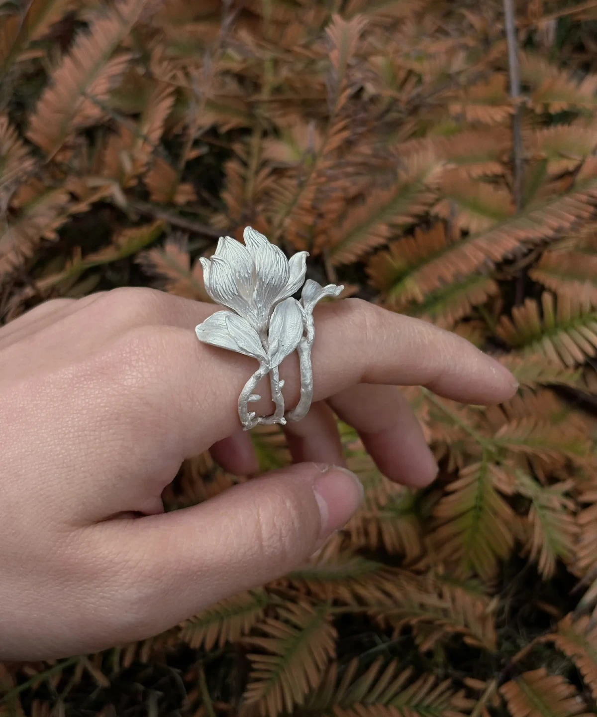 Handmade S925 Sterling Silver Magnolia Ring worn across two fingers, with sculpted petals and stems; a nature-inspired Silver Ring showcasing Magnolia Jewelry craftsmanship against a backdrop of dried fern leaves.