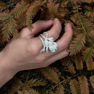 Hand wearing a Handmade S925 Sterling Silver Magnolia Ring with sculpted petals and stem details, resting on fern leaves; a nature-inspired Silver Ring that blends Magnolia Jewelry artistry with botanical elegance.