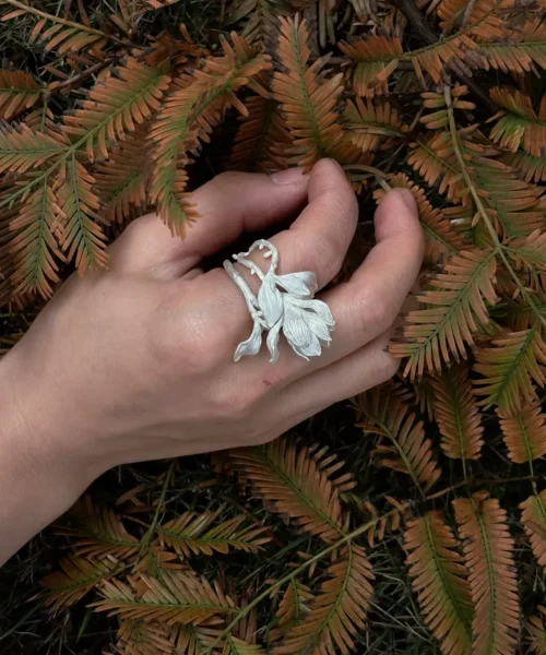 Hand wearing a Handmade S925 Sterling Silver Magnolia Ring with sculpted petals and stem details, resting on fern leaves; a nature-inspired Silver Ring that blends Magnolia Jewelry artistry with botanical elegance.