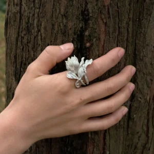Hand wearing a Handmade S925 Sterling Silver Magnolia Ring with sculpted petals and stem details, resting against tree bark; a nature-inspired Silver Ring showcasing Magnolia Jewelry artistry and botanical elegance.