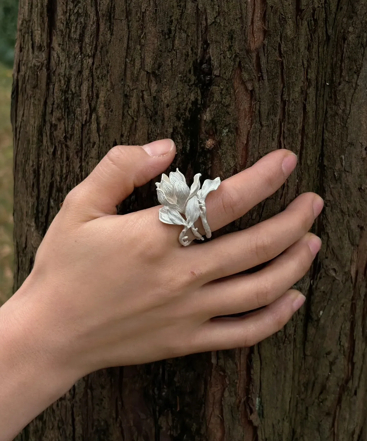 Hand wearing a Handmade S925 Sterling Silver Magnolia Ring with sculpted petals and stem details, resting against tree bark; a nature-inspired Silver Ring showcasing Magnolia Jewelry artistry and botanical elegance.