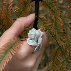 Hand wearing a Handmade S925 Sterling Silver Magnolia Ring with sculpted white petals and stem details, gently holding autumnal leaves; a nature-inspired Silver Ring showcasing Magnolia Jewelry elegance and botanical craftsmanship.
