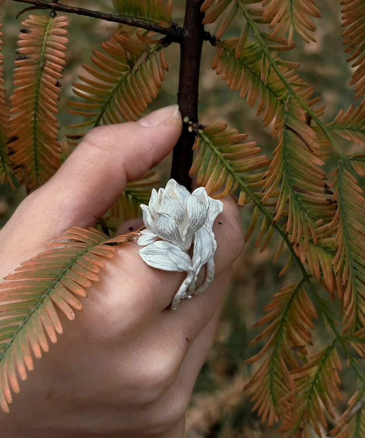 Hand wearing a Handmade S925 Sterling Silver Magnolia Ring with sculpted white petals and stem details, gently holding autumnal leaves; a nature-inspired Silver Ring showcasing Magnolia Jewelry elegance and botanical craftsmanship.