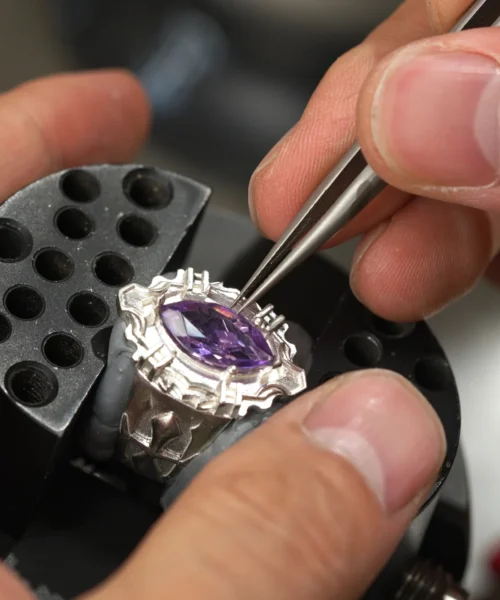 Close-up of a jeweler setting a marquise-cut purple gemstone into a Custom Vongola Silver Ring using precision tweezers; the ring is secured in a clamp, highlighting the craftsmanship and detail of this Handmade Custom Ring in Sterling Silver.