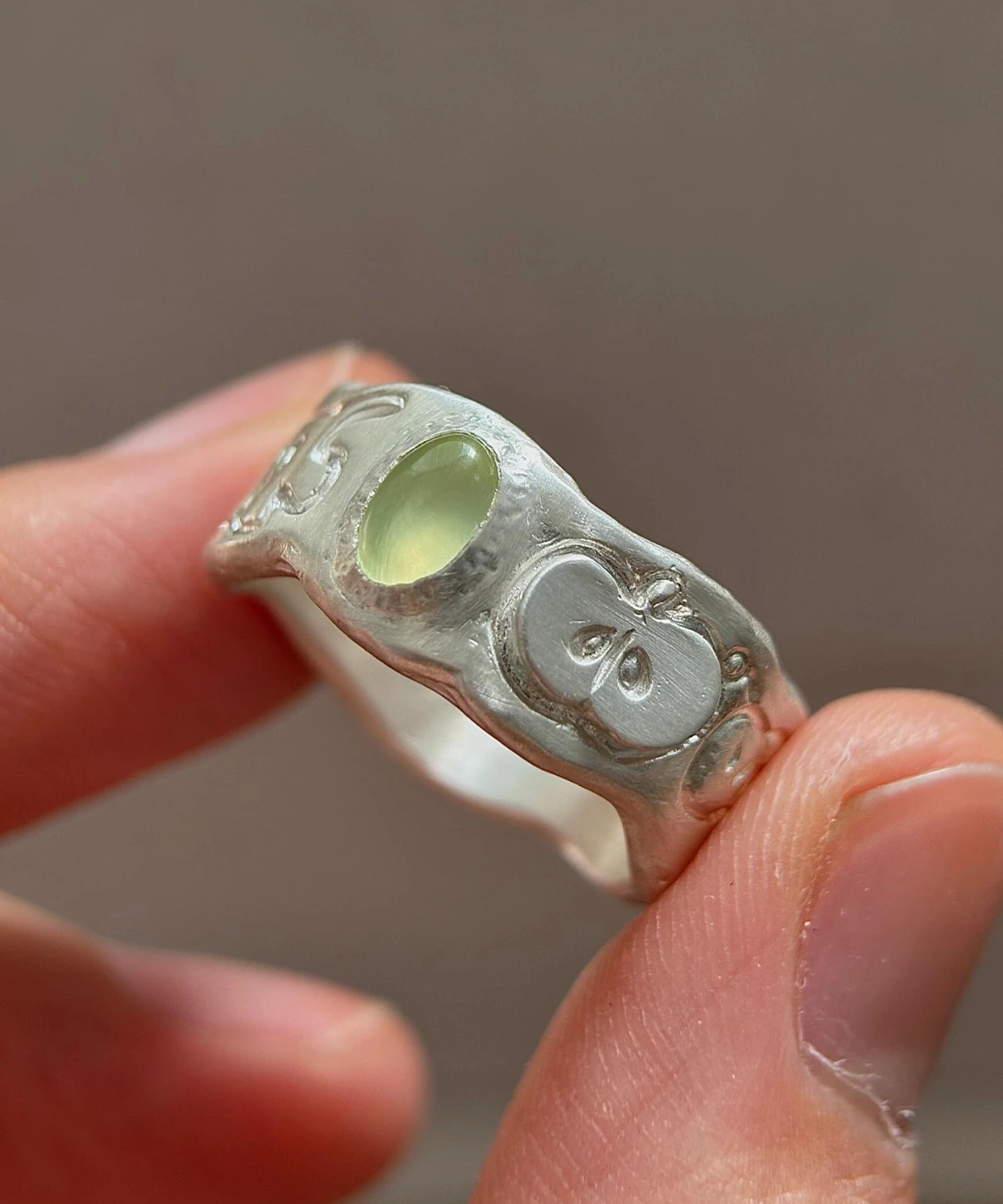 Natural Peridot Ring in 999 Silver with green oval gemstone and engraved apple motif; a handcrafted 999 Silver Ring featuring symbolic leaf and seed details, held between fingers in close-up view.