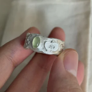 Natural Peridot Ring in 999 Silver featuring green oval gemstone and engraved apple motif with leaf and seeds; a handcrafted 999 Silver Ring held between fingers in close-up view, showcasing symbolic and organic design.
