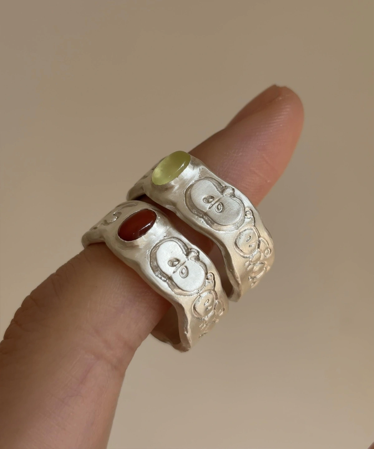 Two handcrafted 999 Silver Rings featuring stylized face engravings; one with a Natural Peridot gemstone in pale green, the other with a red South Red Agate. Unique artisan rings worn on hand in close-up view.
