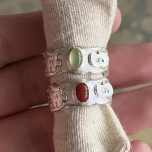 Two handcrafted 999 Silver Rings featuring stylized ghost and pumpkin engravings; one with a pale green Natural Peridot gemstone, the other with a reddish-brown South Red Agate. Unique artisan rings styled on beige fabric in close-up view.