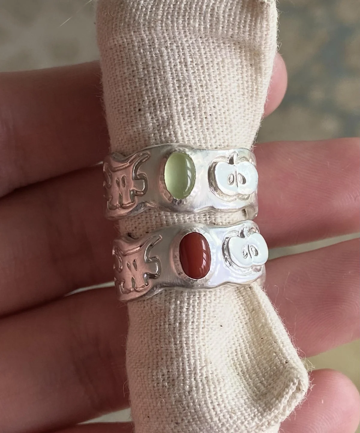 Two handcrafted 999 Silver Rings featuring stylized ghost and pumpkin engravings; one with a pale green Natural Peridot gemstone, the other with a reddish-brown South Red Agate. Unique artisan rings styled on beige fabric in close-up view.