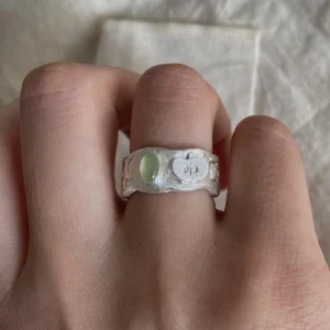 Natural Peridot Ring in 999 Silver featuring light green oval gemstone and engraved apple motif with leaf and seed details; a handcrafted 999 Silver Ring with textured band, worn on hand against soft beige fabric.