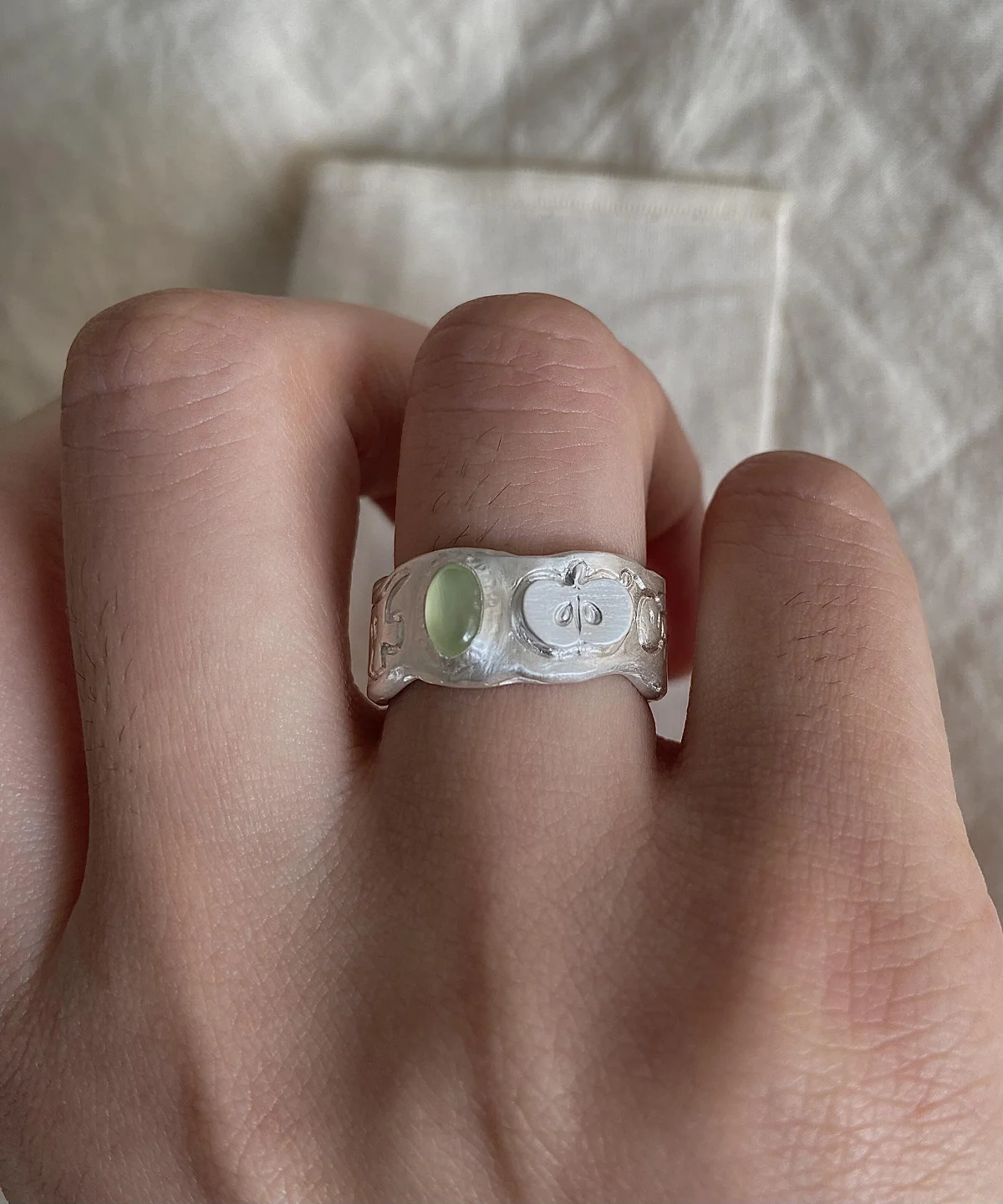 Natural Peridot Ring in 999 Silver featuring light green oval gemstone and engraved apple motif with leaf and seed details; a handcrafted 999 Silver Ring with textured band, worn on hand against soft beige fabric.