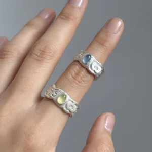 Two handcrafted 999 Silver Rings featuring engraved apple motifs and oval gemstones—one pale green Natural Peridot Ring and one soft blue Natural Aquamarine Ring—worn on adjacent fingers in close-up view.