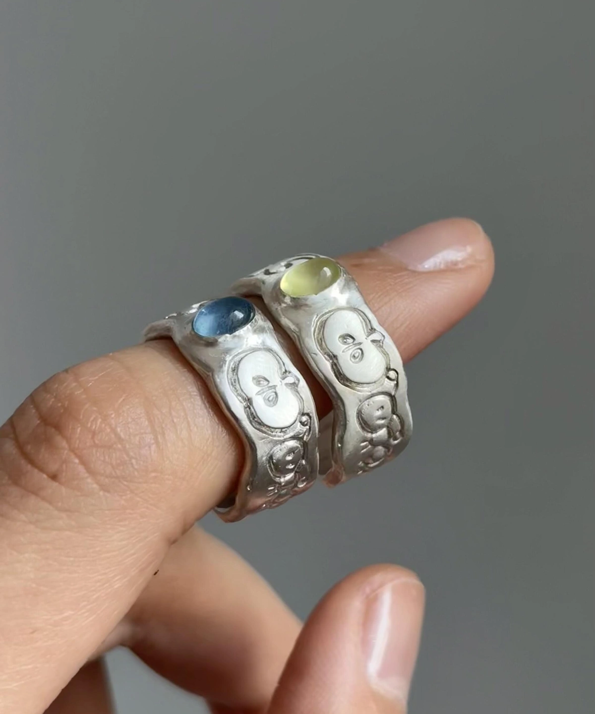 Two handcrafted 999 Silver Rings featuring stylized face motifs and central oval gemstones—one pale green Natural Peridot Ring and one soft blue Natural Aquamarine Ring—worn on adjacent fingers in close-up view.