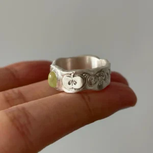 Natural Peridot Ring in 999 Silver with green oval gemstone and engraved apple motif; a handcrafted 999 Silver Ring featuring symbolic leaf and seed details, held between fingers in close-up view.