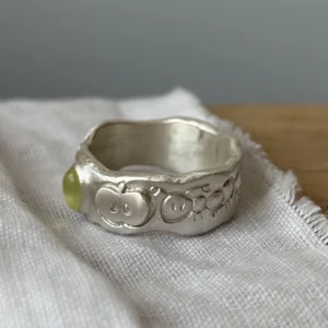 Natural Peridot Ring in 999 Silver featuring light green oval gemstone and engraved stick-figure apple face motifs; a handcrafted 999 Silver Ring with organic texture and whimsical design, styled on fabric in close-up view.