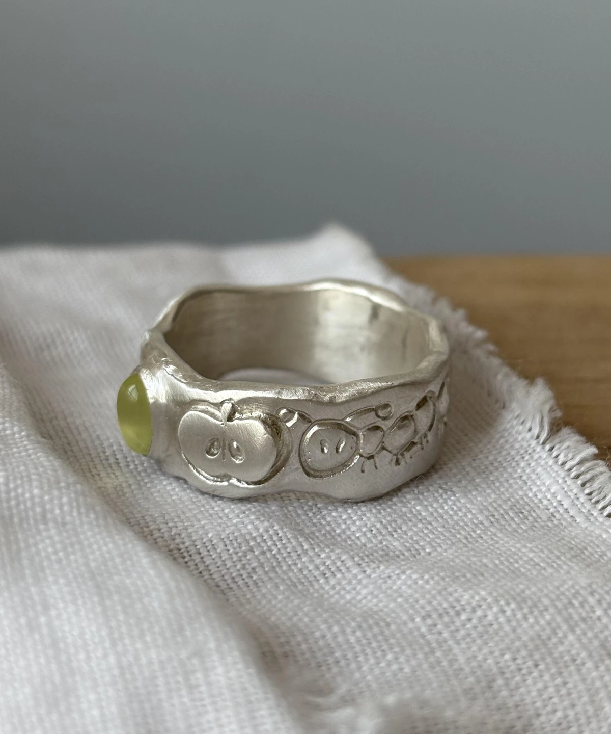 Natural Peridot Ring in 999 Silver featuring light green oval gemstone and engraved stick-figure apple face motifs; a handcrafted 999 Silver Ring with organic texture and whimsical design, styled on fabric in close-up view.