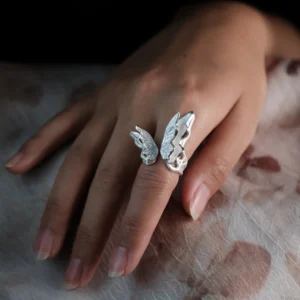Close-up of a hand wearing a S925 Sterling Silver Butterfly Ring with lifelike textured wings in an open wrap design, resting on a floral-patterned surface; elegant Silver Ring showcasing handcrafted detail and nature-inspired Butterfly Ring artistry.
