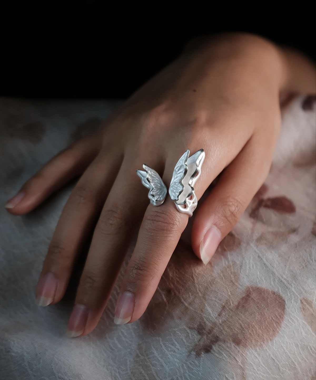 Close-up of a hand wearing a S925 Sterling Silver Butterfly Ring with lifelike textured wings in an open wrap design, resting on a floral-patterned surface; elegant Silver Ring showcasing handcrafted detail and nature-inspired Butterfly Ring artistry.