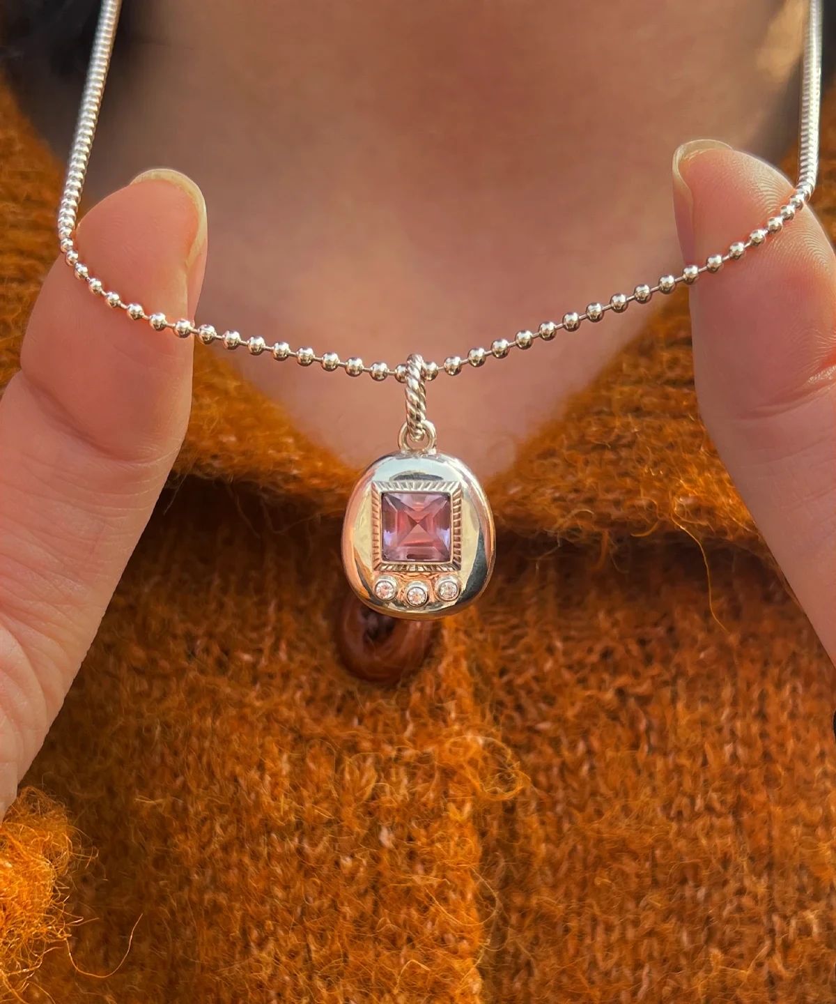 Custom Handmade S925 Sterling Silver Tamagotchi Necklace 002 Tamagotchi Necklace with light purple gemstone and three clear accent stones in S925 silver; held by hands over rust-colored fabric, showcasing Pet Necklace craftsmanship and nostalgic elegance.