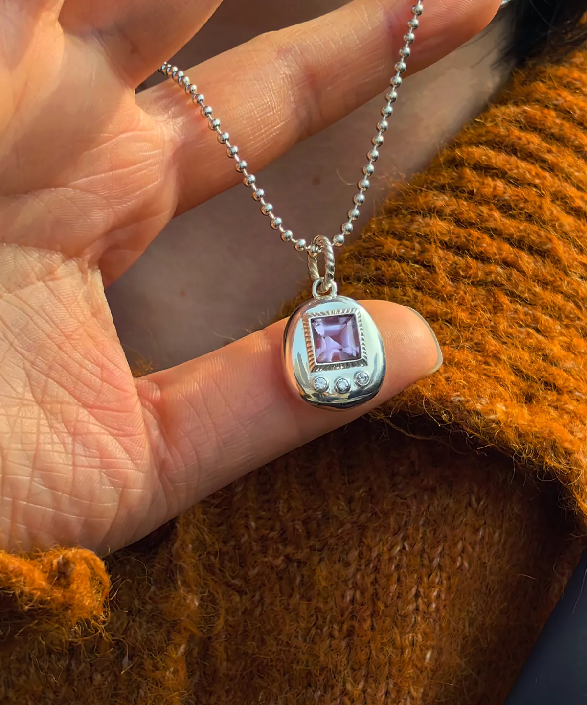 Custom Handmade S925 Sterling Silver Tamagotchi Necklace 04 Tamagotchi Necklace with light purple square gemstone and three clear accent stones in S925 silver, held by hand over rust-colored fabric.