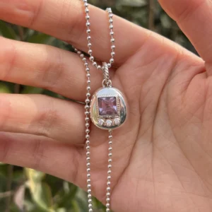 Tamagotchi Necklace with light purple square gemstone and three clear accent stones in S925 silver; held by hand outdoors with green foliage background, showcasing Pet Necklace craftsmanship and nostalgic charm.
