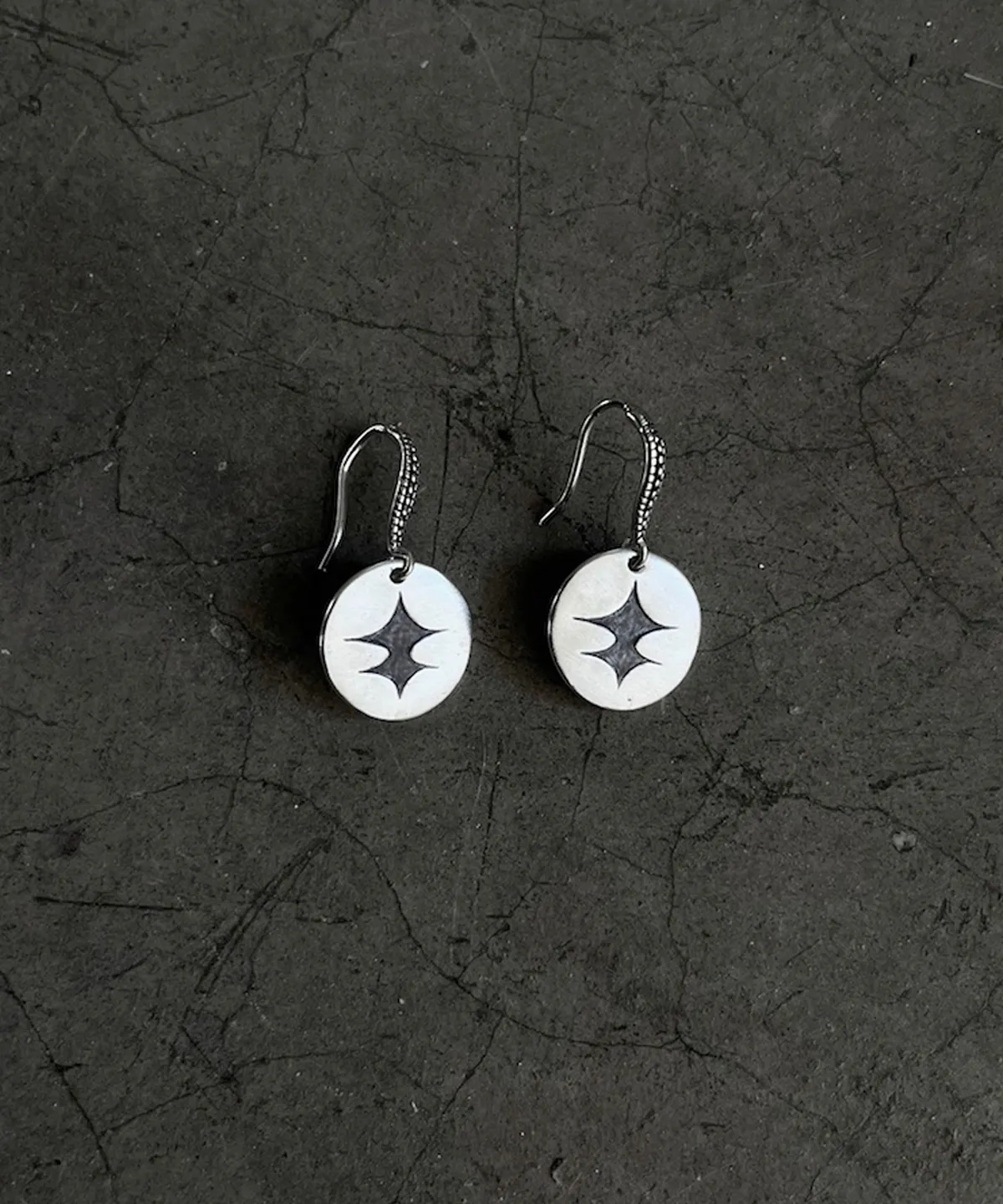 Disc Earrings in S925 Sterling Silver with engraved black starburst design and textured hooks; placed on cracked dark surface to highlight handcrafted detail, modern contrast, and artistic elegance in engraved earrings.