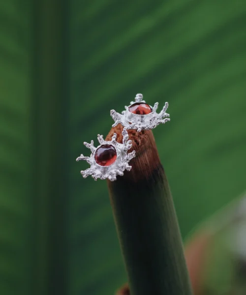 Natural Garnet Stud Earrings in S925 Sterling Silver with splash-inspired silverwork and reddish-brown gemstones; styled on plant stem tip against green leaf backdrop to highlight organic elegance and handcrafted artistry.