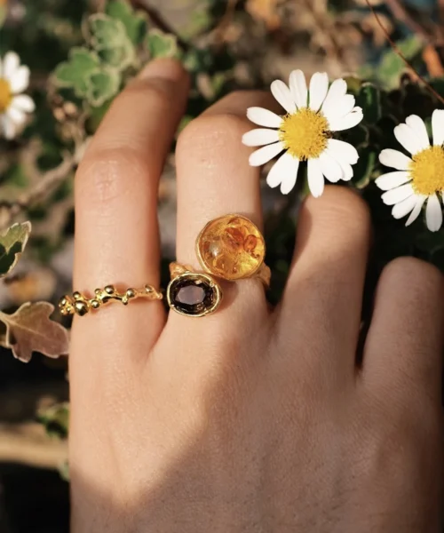 Hand wearing three gold rings, including an Amber Ring with warm golden and deep purple gemstones, shown among white daisies and green foliage.